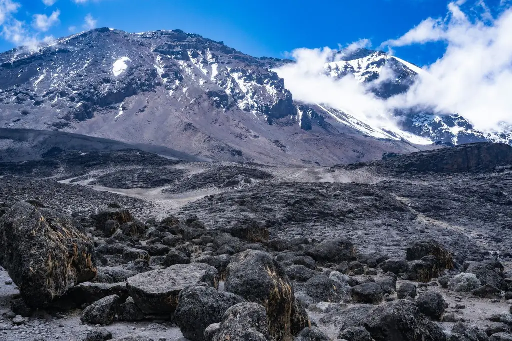 A snow-streaked summit cone above a field of dark volcanic boulders, cloud drifting past